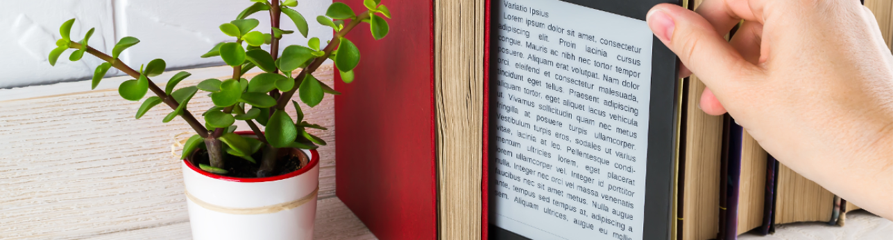 A person's hand holding an e-reader displaying text, surrounded by books and a small potted plant with green leaves, against a wooden surface.