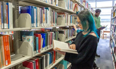 OER event with David Harris, editor of OpenStax A woman with blue hair examines a book while seated in a library, surrounded by shelves filled with books