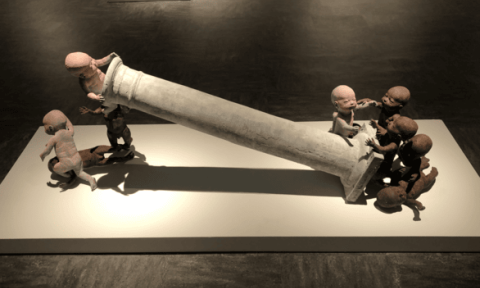 Art installation of babies pulling a toppled column, each straining with effort. The dim lighting and their focused expressions convey determination.