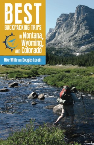 A hiker with a backpack crosses a rocky stream in a lush, green mountainous landscape. A towering cliff rises under a clear blue sky. Text overlay reads: "Best Backpacking Trips in Montana, Wyoming, and Colorado" by Mike White and Douglas Lorain.