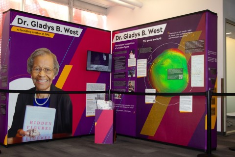 New Library Exhibit Highlights Unsung Mathematician Exhibit honoring Dr. Gladys B. West, featuring biography, a book titled "Hidden Figures," and a colorful geoid model. The tone is informative and celebratory.