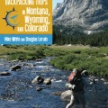 A hiker with a backpack crosses a rocky stream in a lush, green mountainous landscape. A towering cliff rises under a clear blue sky. Text overlay reads: "Best Backpacking Trips in Montana, Wyoming, and Colorado" by Mike White and Douglas Lorain.