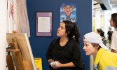 Two young women closely observe artwork displayed on a wall in an indoor setting, with other people and art visible in the background.