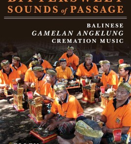 Group of Balinese musicians in orange shirts and traditional headwear play Gamelan Angklung instruments in a shaded outdoor setting, conveying cultural vibrancy.